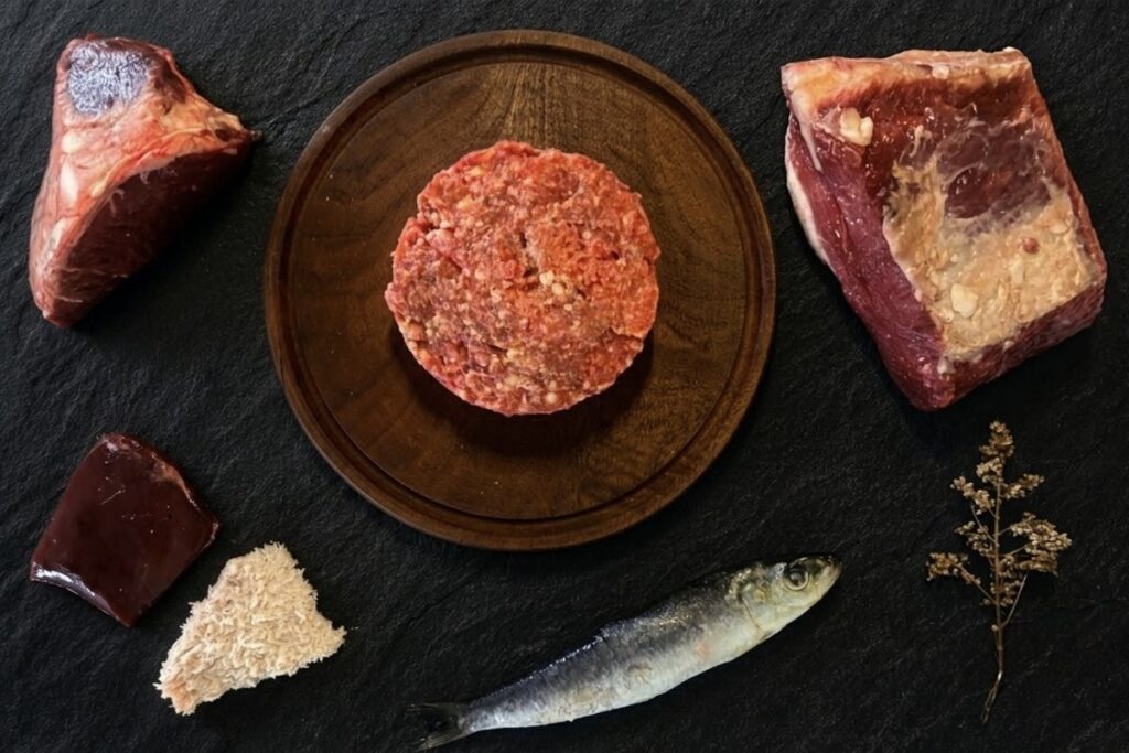 Menú BARF de carne y pescado para gatos: hamburguesa de alimento crudo sobre tabla de madera, rodeada de un corte de carne vacuna, sardina entera, hígado, hueso carnoso y mondongo, sobre pizarra negra