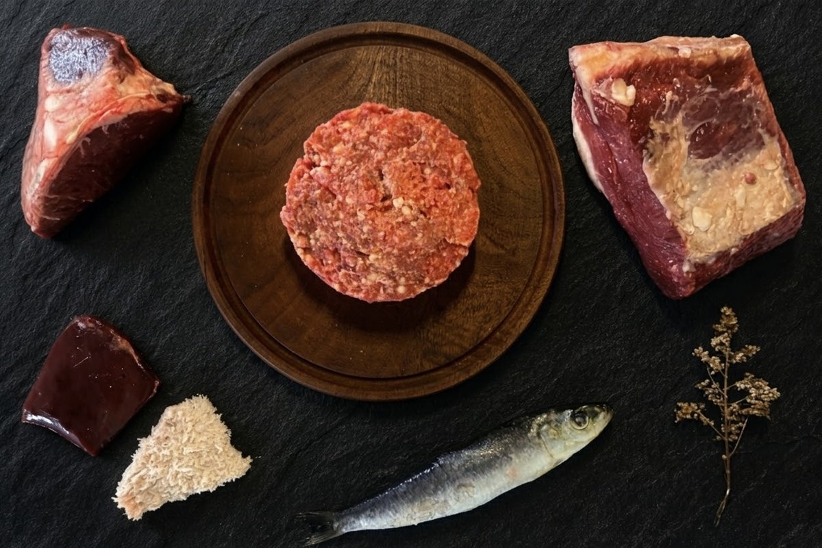 Menú BARF de carne y pescado para gatos: hamburguesa de alimento crudo sobre tabla de madera, rodeada de un corte de carne vacuna, sardina entera, hígado, hueso carnoso y mondongo, sobre pizarra negra