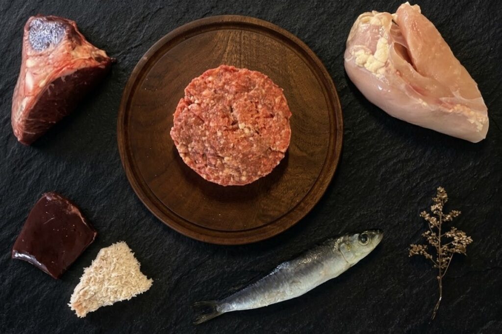 Menú BARF de pollo y pescado para gatos: hamburguesa de alimento crudo sobre tabla de madera, rodeada de pechuga de pollo, sardina entera, hígado, hueso carnoso y mondongo, sobre pizarra negra