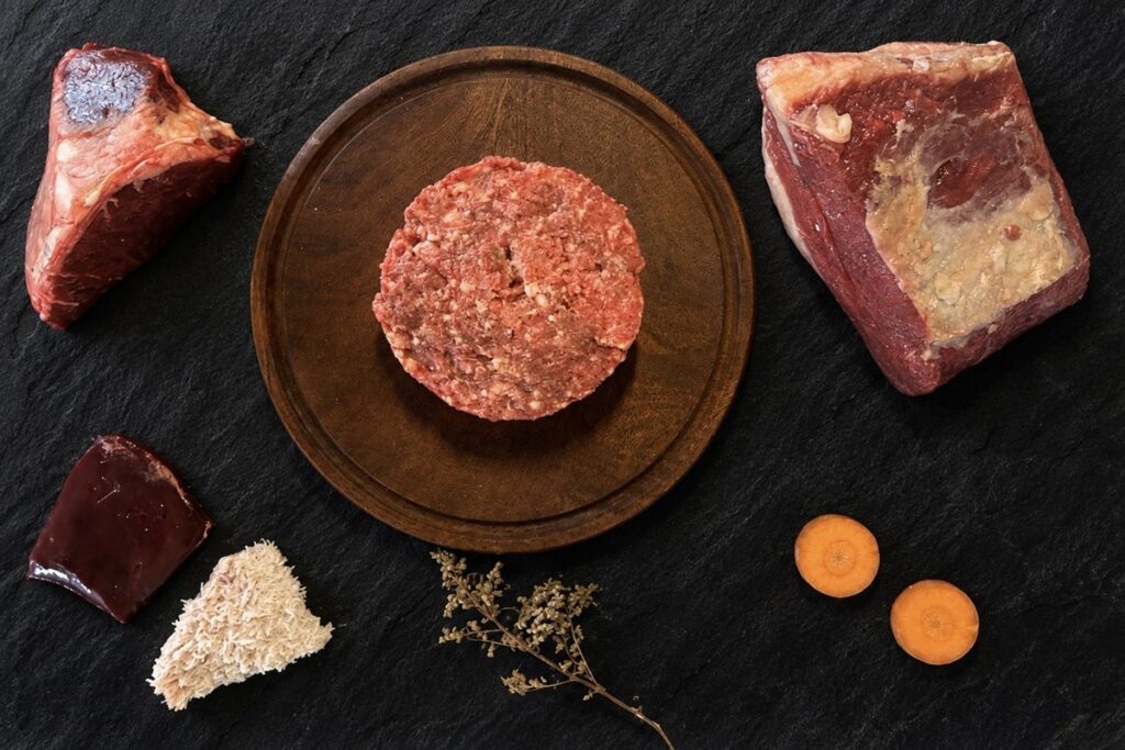 Menú BARF de carne para perros: hamburguesa de alimento crudo sobre tabla de madera, rodeada de dos cortes de carne vacuna, hígado, mondongo y rodajas de zanahoria, sobre pizarra negra