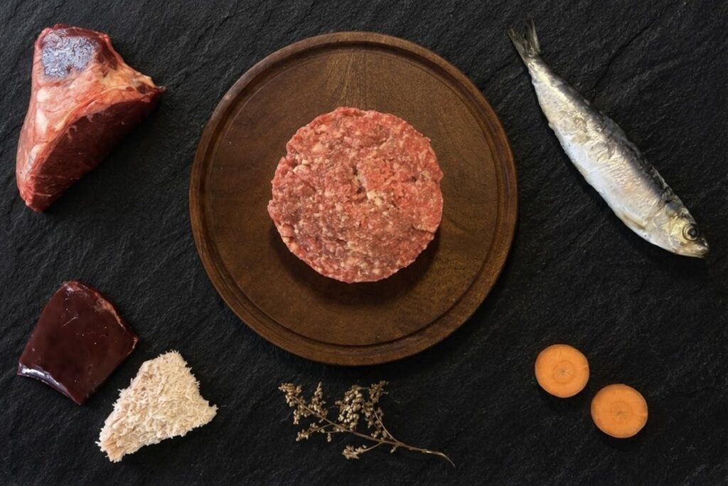 Menú BARF de pescado para perros: hamburguesa de alimento crudo sobre tabla de madera, rodeada de una sardina entera, hígado, hueso carnoso, mondongo y rodajas de zanahoria, sobre pizarra negra