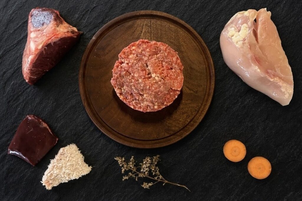Menú BARF de pollo para perros: hamburguesa de alimento crudo sobre tabla de madera, rodeada de pechuga de pollo, hígado, hueso carnoso, mondongo y rodajas de zanahoria, sobre pizarra negra